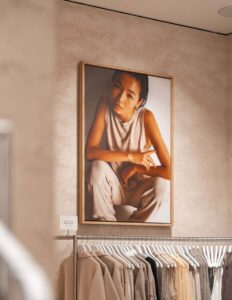 Store interior with large portrait of woman above rack of beige clothing on hangers.