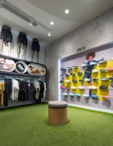 Children's athletic shoe display in retail store with mannequins, clothing racks, and seating on green carpet.