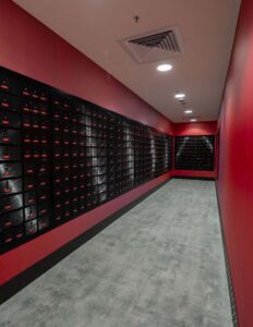 Red hallway with rows of black mail lockers on both sides, grey flooring, overhead lights, modern postal storage design.