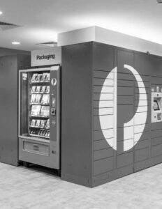 Vending machine with packaging area in modern lobby, featuring lockers and sleek design.
