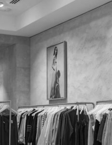 Clothing store interior with racks of assorted garments and a framed black-and-white fashion photo on the wall.