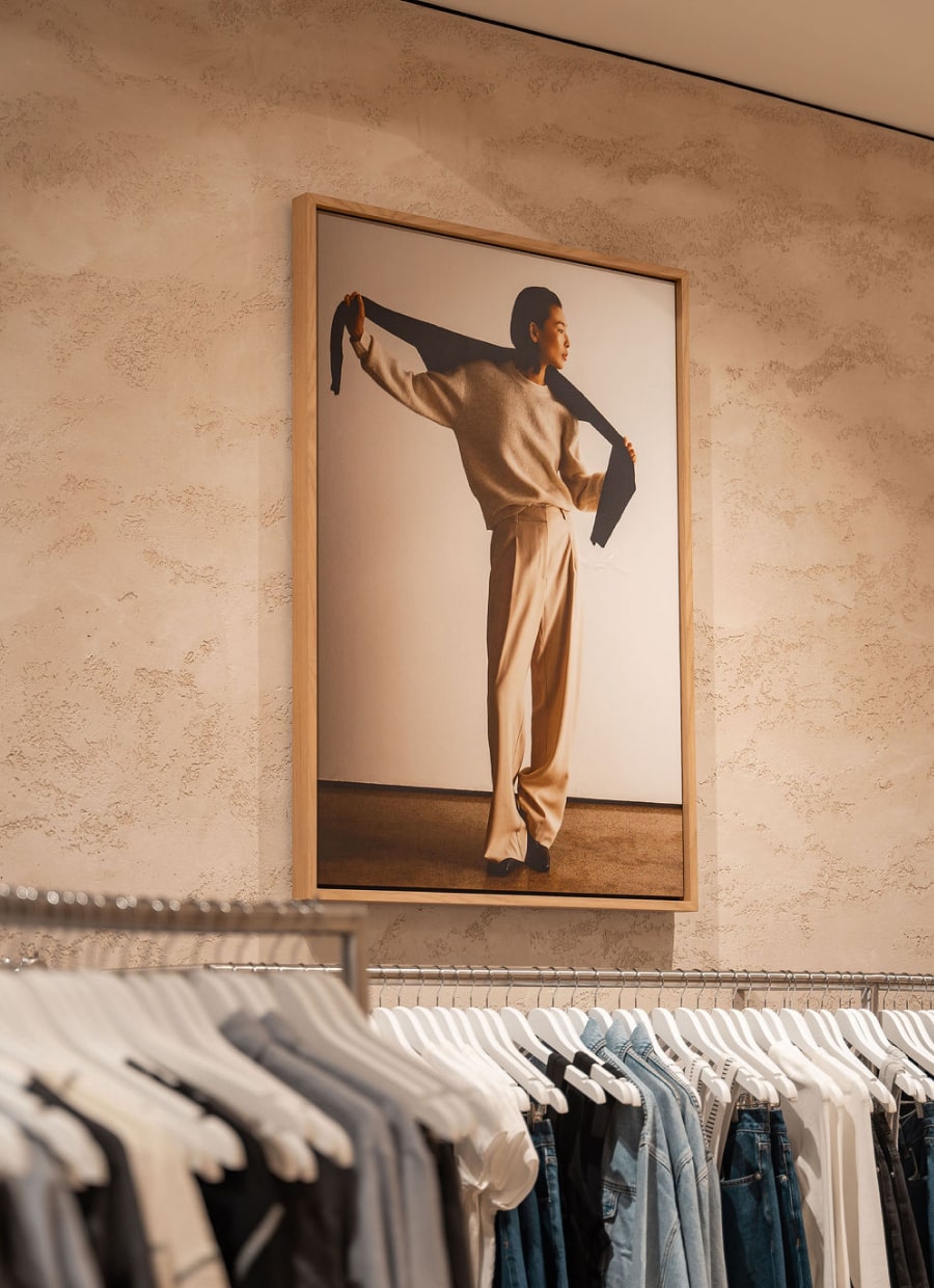 Fashion store interior with large framed photo of a model in beige outfit, above clothing rack with shirts and jackets.