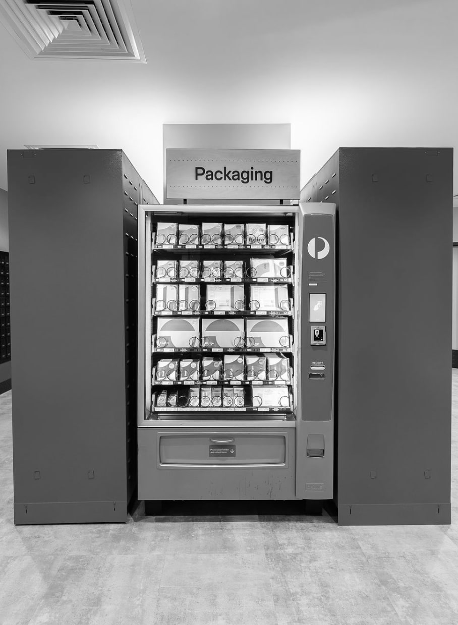 Vending machine for packaging supplies, black-and-white photo, offering various packing materials, indoor setup.