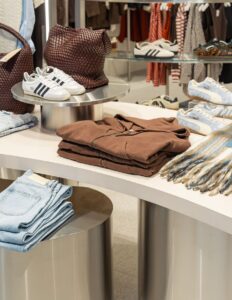 Clothing store display with sneakers, woven bag, brown hoodie, denim jeans, and colorful scarf on modern tables.