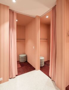Modern dressing room with pink walls, curtains, and soft lighting, featuring plush seating and muted tones.